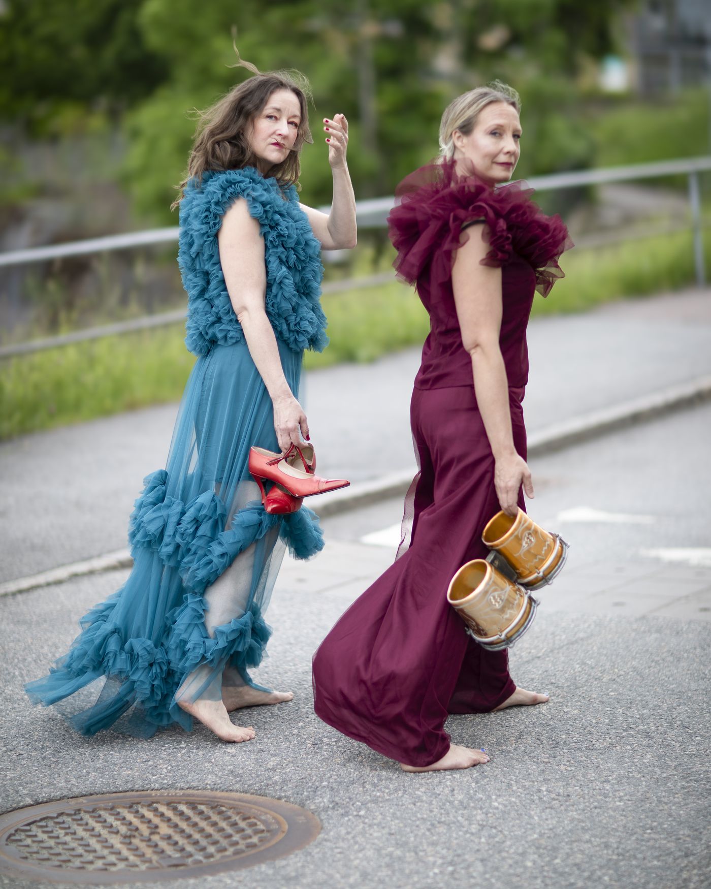 Julia and Yohanna walking barefoot along a road in blue and wine-red tulle dresses, shoes in hand